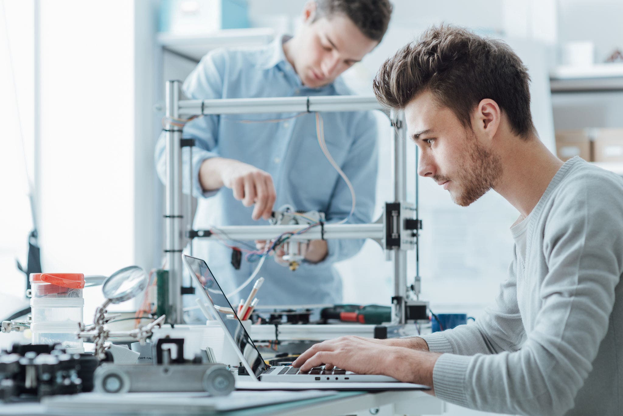 Engineering students working in the lab, a student is adjusting a 3D printer's components, the other one on foreground is using a laptop