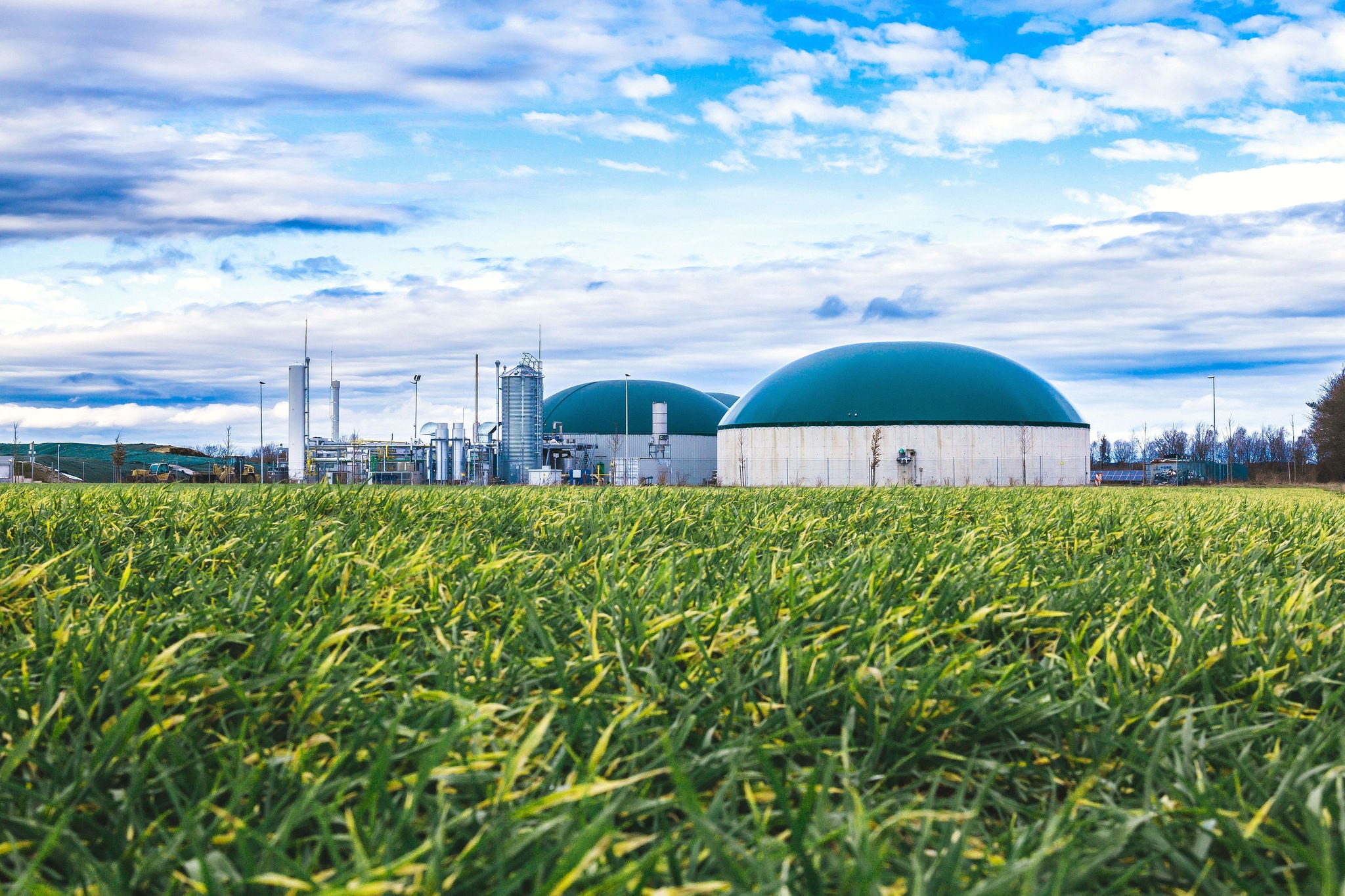 Renewable Energy Biogas Biogas plant with green storage domes situated in a lush grassy field under a partly cloudy blue sky.