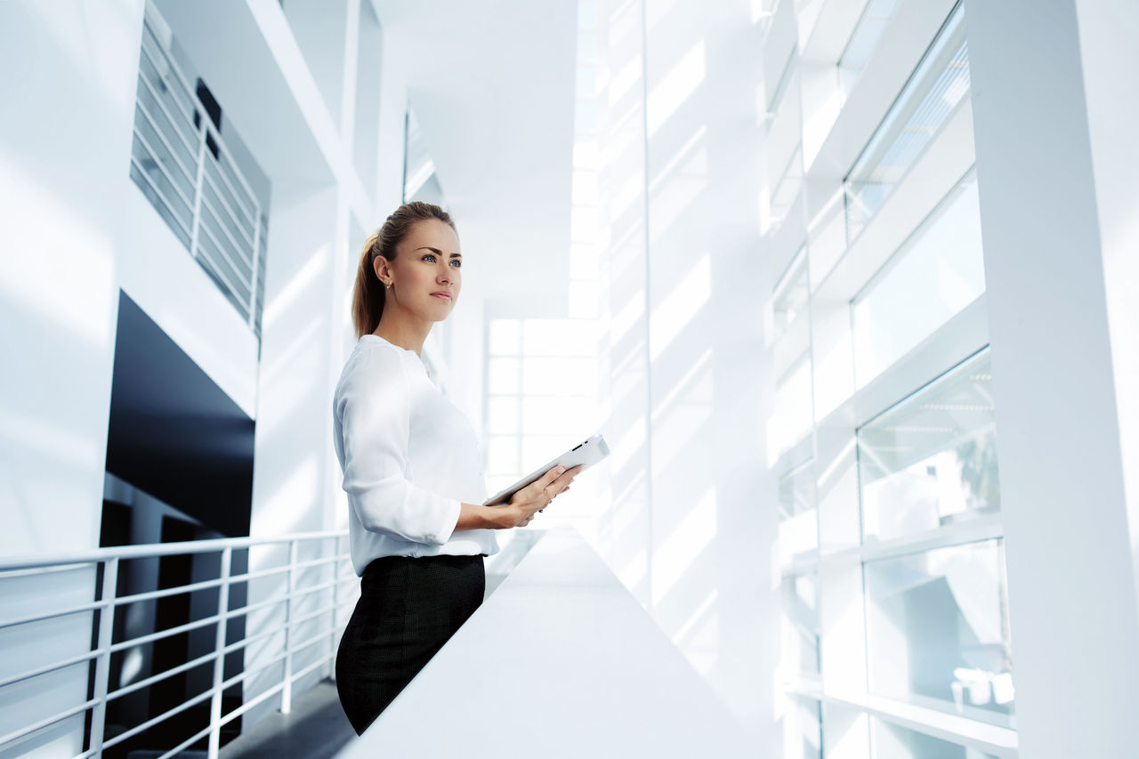 Thoughtful woman holding digital tablet and looks into the window after business meeting with partners, young successful female financier ponders over the future of company during work break in office